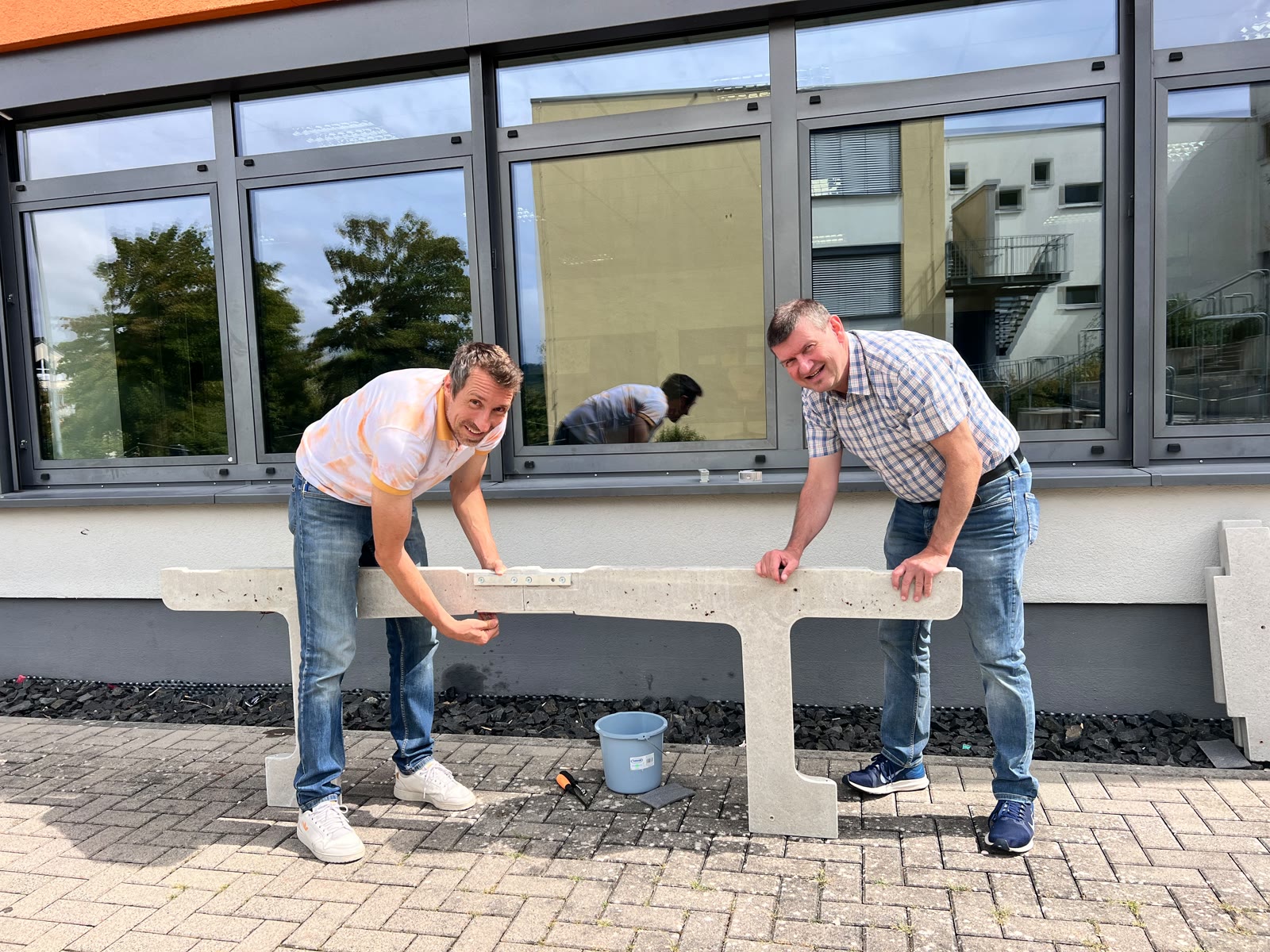 Zwei Lehrkräfte montieren ein Betonelement der neuen Tischtennisplatte im Außenbereich der WKS Weilburg.